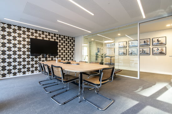 Salle de réunion qui accueille une table de réunion et huit chaises noires Marcel Breuer. Un mur est décoré d'!un papier peint vintage et au milieu est disposé un grand écran télé.